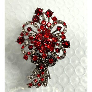 May include: A silver-toned brooch in the shape of a floral bouquet. The brooch is adorned with numerous red gemstones of varying sizes, creating a sparkling effect. The design includes bow-shaped elements and detailed metalwork.