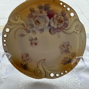 May include: A vintage porcelain plate with a floral rose design. The plate features a cream-colored center with hand-painted roses in shades of white, pink, and purple. The rim is a golden-yellow color with decorative cut-out holes.