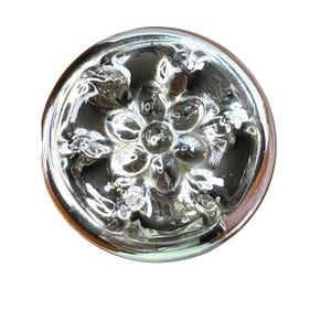 May include: A clear, round glass bottle stopper with a floral design. The stopper has a raised, detailed pattern of flowers and leaves. The glass has a reflective, shiny surface, and the stopper is set against a white background.