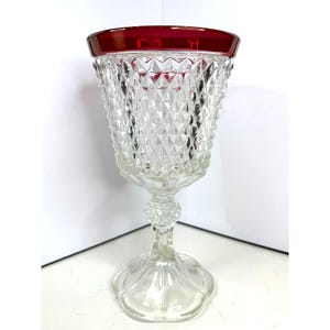 May include: A clear glass goblet with a diamond pattern and a red rim. The goblet has a tall stem and a wide base.