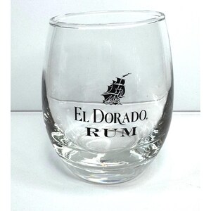 May include: A clear, stemless glass tumbler featuring a black graphic of a sailing ship above the black printed text EL DORADO RUM. The glass has a rounded, bulbous shape with a thick base and a smooth, transparent finish throughout the body.