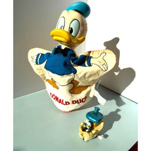 May include: A vintage Donald Duck puppet with a blue hat, shirt, and bow tie. The puppet has a yellow beak and is made of fabric and plastic. A small Donald Duck toy is also present.