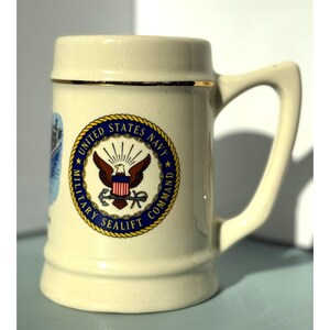 May include: Cream-colored ceramic beer stein with a handle and gold trim. The stein features a circular emblem with the words "United States Navy Military Sealift Command" and an eagle.