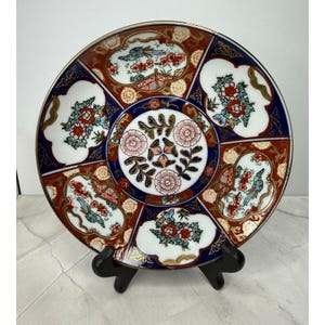 May include: Decorative ceramic plate with a gold rim, featuring a detailed floral and bird design. The plate has a white, red, and blue color scheme with intricate patterns. The plate is displayed on a black stand.