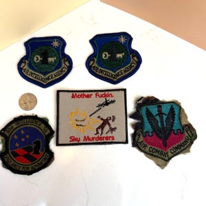 May include: A collection of embroidered patches with military-themed designs. The patches feature text such as "Air Intelligence Agency", "938 Intelligence Sq", and "Air Combat Command". One patch has the text "Mother Fudsin" and "Sky Murderers".