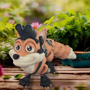 May include: A plush fox toy with a cartoonish design. The fox has orange fur, a white muzzle, and black accents on its ears, paws, and a headpiece. It is posed on a wooden surface with a blurred garden background.