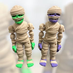 May include: Two articulated mummy figurines, one with green and one with purple painted details. The figures are made of beige plastic and have a detailed mummy design.