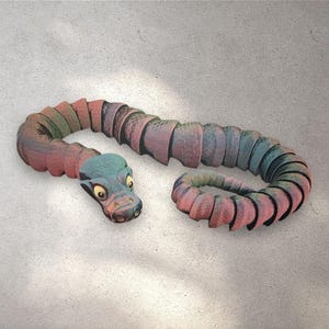 Puede incluir: Una escultura de serpiente segmentada y caprichosa en tonos rojos, verdes y azules. La cabeza de la serpiente es de color turquesa con ojos amarillos. La escultura está sobre una superficie gris claro texturizada.
