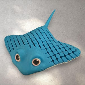 May include: A blue stingray sculpture with a textured, grid-like pattern on its wings. The sculpture has large, round eyes with pink accents and a pointed tail. The background is a light gray, textured surface.
