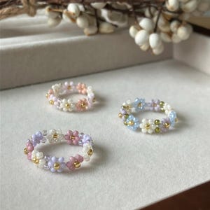May include: Three beaded rings in pastel colors. Each ring features a floral design with small beads in shades of pink, white, purple, and blue. Gold-colored beads accent the flowers. The rings are displayed on a neutral surface.