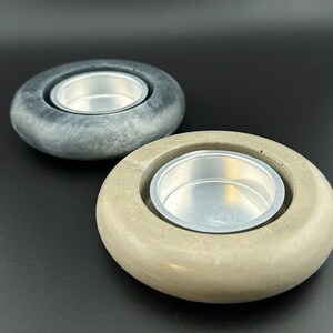 May include: Two round concrete candle holders with a silver metal insert. The holder on the left is dark gray and the holder on the right is light brown.