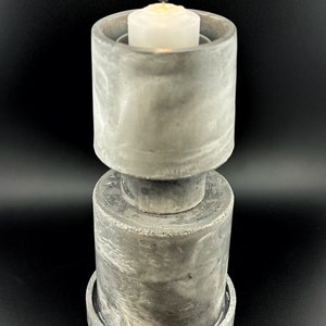 May include: A gray concrete candle holder with a white candle inside. The candle holder has a cylindrical shape with a wider base and a smaller top. The candle is lit and the flame is visible.