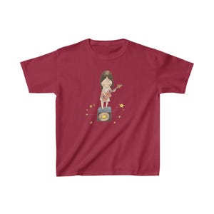 May include: A red youth-sized t-shirt with a cartoon illustration of a girl playing a guitar on top of an amplifier. The girl is wearing a pink dress and has brown hair. There are yellow stars around the girl and amplifier.