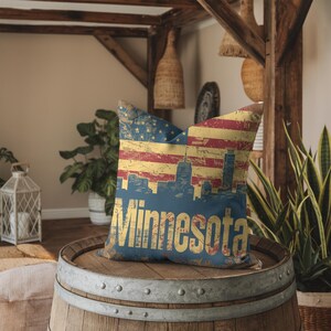 Puede incluir: Una almohada decorativa con un diseño de bandera estadounidense desgastada y la palabra "Minnesota" en una fuente vintage. La almohada está sentada sobre un barril de madera.