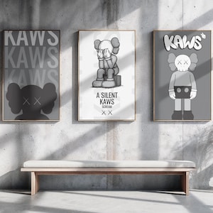 May include: Three framed KAWS art prints in a modern interior. The prints feature the KAWS character in various poses and designs, with text including "KAWS" and "A Silent KAWS". The color palette is primarily grayscale with a wooden bench and a vase with dried plants.
