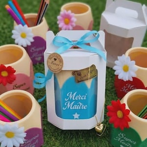 Peut inclure: Plusieurs pots à crayons personnalisés, certains avec des crayons, et une boîte cadeau avec un ruban bleu. La boîte cadeau porte l'inscription "Merci Maître". Chaque pot est décoré d'une fleur.
