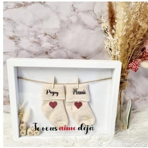 Op de afbeelding: Een wit fotolijstje met een witte achtergrond. Binnen het lijstje hangen twee paar babysokken aan een waslijn. De sokken zijn gelabeld met "Papy" en "Mamie". Onder de sokken zijn er houten blokken die "BABY" spellen. De tekst "Je vous aime deja" staat onder de blokken.