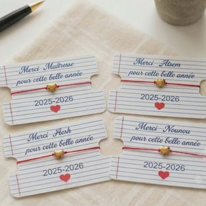Peut inclure: Quatre bracelets en fil rouge avec un petit cœur doré, montés sur des cartes rectangulaires imitant du papier ligné. Les cartes portent les inscriptions Merci Maîtresse, Atsem, Aesh ou Nounou pour cette belle année, 2025-2026, et une icône de cœur rouge.