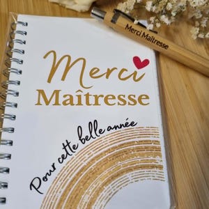 Peut inclure: Un carnet à spirales blanc avec le texte doré Merci Maîtresse et un cœur rouge, ainsi que le texte Pour cette belle année au-dessus d'un motif en demi-cercle doré. Un stylo en bambou assorti avec le texte Merci Maîtresse est posé dessus.
