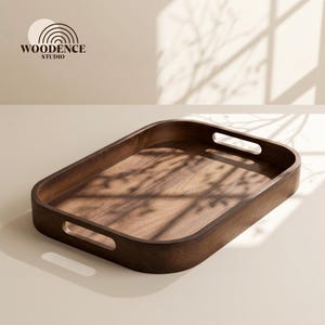 Puede incluir: Una bandeja de servicio rectangular de madera marrón oscuro con esquinas redondeadas y dos asas recortadas integradas en los lados cortos. La bandeja presenta una superficie lisa y pulida con vetas de madera visibles y el texto WOODENCE STUDIO.