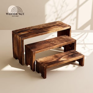 May include: A set of three tiered wooden display risers with a dark, scorched wood grain finish. The rectangular stands are arranged in a cascading formation, each featuring a flat top surface and two side supports. The logo Woodence Studio is visible.