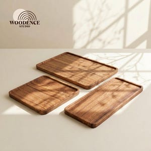 Op de afbeelding: Drie rechthoekige houten serveerbladen in verschillende maten. De bladen zijn gemaakt van donker hout met zichtbare nerfpatronen. Ze zijn gerangschikt op een licht oppervlak. Het logo "WOODENCE STUDIO" is linksboven zichtbaar.