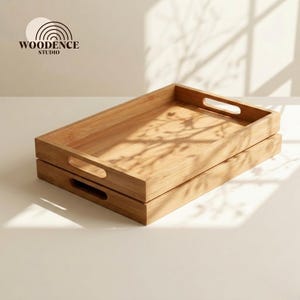 May include: Two rectangular wooden serving trays stacked. Each tray has a cutout handle on each side. The trays are made of light-coloured wood and are on a white surface. The logo "WOODENCE STUDIO" is in the upper left corner.