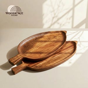 Op de afbeelding: Twee houten serveerschalen met een bladachtige vorm. De schalen zijn gemaakt van warm getint hout en hebben een handvat. Het logo "WOODENCE STUDIO" is zichtbaar in de linkerbovenhoek. De schalen zijn op elkaar gestapeld.