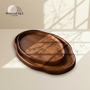 May include: Two oval wooden serving trays nested together. The trays are made of dark brown wood with a smooth finish. The trays are on a light beige surface, with the logo "WOODENCE STUDIO" in the upper left corner.