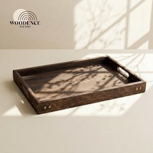 Op de afbeelding: Een rechthoekige houten serveerschaal met een donkerbruine afwerking. De schaal heeft een opstaande rand, uitgesneden handgrepen en messingkleurige klinknagels. Het Woodence Studio-logo staat linksboven.
