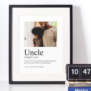 Può includere: Stampa incorniciata con la definizione di "Zio" e una foto di un uomo e un bambino. La cornice nera circonda un passe-partout bianco e la stampa. Un orologio a scatto nero e due libri sono su una superficie bianca.