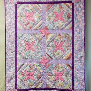 May include: Handmade quilt featuring a central design of four star-shaped blocks in shades of pink and blue. The stars are set against a patchwork background with a lavender border and floral patterned edges. The quilt is a cozy, decorative textile.