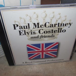 Puede incluir: Caja de CD con el texto "Paul McCartney Elvis Costello and friends" encima de una bandera de la Union Jack. La caja es de plástico transparente, con fondo blanco y texto negro. El texto "A Royal Command Performance" está en la parte inferior.