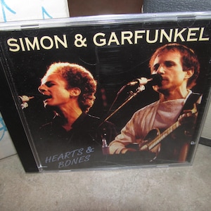 May include: A Simon & Garfunkel music CD titled Hearts & Bones. The cover features a black background with the band name in white text at the top and the album title in blue text at the bottom. Two musicians are shown singing into microphones.