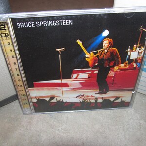 Puede incluir: Una caja de CD con Bruce Springsteen en el escenario, sosteniendo una guitarra. La portada muestra a Springsteen con una chaqueta marrón y pantalones negros, con un coche rojo en el fondo. El texto "BRUCE SPRINGSTEEN" está en la parte superior.
