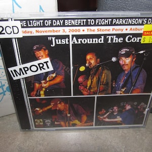 May include: A 2CD album cover with a black background and white text. The album title is "Just Around The Corner". The album cover features four musicians, three of whom are playing guitars. The album is a benefit for Parkinson's Disease. The album was released on 3 November 2000, at The Stone Pony in Asbury Park, New Jersey.