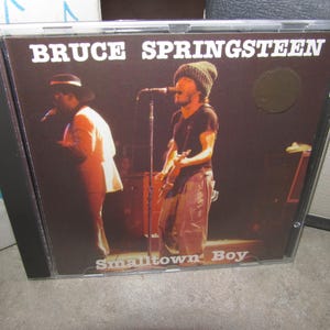 Może przedstawiać: Pudełko na płytę CD z albumem Bruce'a Springsteena. Okładka przedstawia zdjęcie z występu na żywo Springsteena grającego na gitarze, z napisem "BRUCE SPRINGSTEEN" u góry i "Smalltown Boy" na dole. Obraz jest w przezroczystym plastikowym etui.