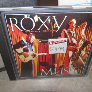 Op de afbeelding: Een cd-hoesje met het album "First Kiss" van Roxy. De cover toont een band die op het podium optreedt met een gitarist en een saxofonist. Het woord "MUSIC" is onderaan zichtbaar.