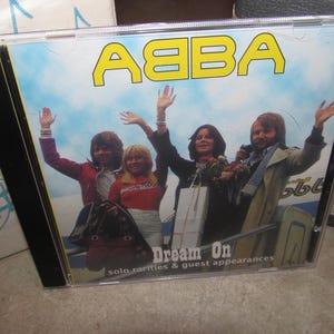 Puede incluir: Una caja de disco compacto con el logotipo de ABBA en amarillo en la parte superior. La portada muestra a cuatro personas saludando con la mano sobre un cielo azul. El texto Dream On solo rarities & guest appearances está en blanco.