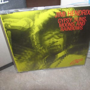 Op de afbeelding: Een cd-hoesje met Jimi Hendrix. De albumhoes is geel met een portret van Hendrix en de tekst "Jimi Hendrix Gypsy Suns, Moons and Rainbows" in het rood. De hoes zit in een doorzichtige plastic hoes.