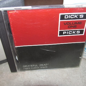 Può includere: Una custodia per CD con "Dick's Volume One Picks" con un design rosso e nero. La custodia del CD è trasparente e la copertina include il testo "Grateful Dead Tampa Florida 12/19/75".
