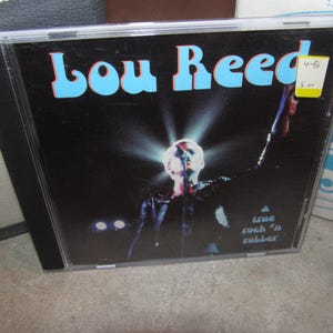 Peut inclure: Un boîtier de CD avec l'album "Lou Reed", le nom de l'artiste en lettres bleu clair. La couverture montre une performance sur scène avec un projecteur sur le chanteur. Le texte "a true rock 'n rubber" est également visible.