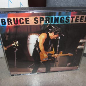 Puede incluir: Una caja de CD con una foto de Bruce Springsteen actuando en un escenario. Lleva una camisa negra y toca la guitarra. El fondo es una imagen borrosa de un escenario de concierto.