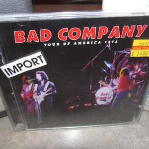 May include: A CD case featuring the album cover for Bad Company's "Tour of America 1974." The cover is black with the band's name in red and a photo of the band performing live. An "IMPORT" sticker is on the case.