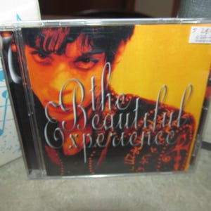 May include: A CD case with a yellow background and a close-up of a person's face. The text on the case reads "The Beautiful Experience".