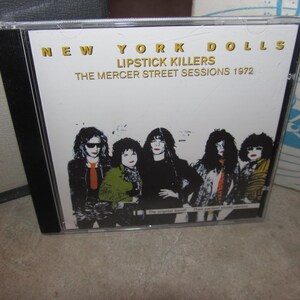May include: A CD case featuring the album "Lipstick Killers: The Mercer Street Sessions 1972" by the New York Dolls. The cover art displays a black and white image of the band members. The text is in gold.