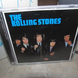 May include: A CD case featuring the band The Rolling Stones. The album cover has a black background with the band's name in blue block letters. The band members are in the foreground.