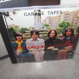 May include: A CD case with the text "GARAGE TAPES" and "FOR SALE BY AUCTION." The cover features a photograph of four men sitting on a bench in front of a building. The men are wearing various outfits, including a yellow shirt and a pink suit.
