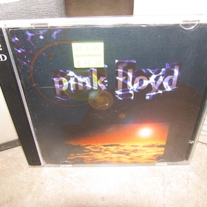 May include: A Pink Floyd 2 CD album in a clear plastic case. The album cover features the band's name in a stylized font over a sunset scene. The case has a sticker with text.