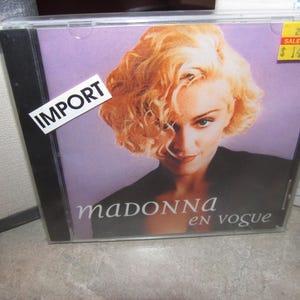 May include: A CD case featuring a portrait of a woman with blonde curly hair. The album cover has the text "madonna en vogue" in white script. A yellow sticker with the text "SALE PRICE $14.99" is on the upper right corner. An "IMPORT" sticker is on the upper left.